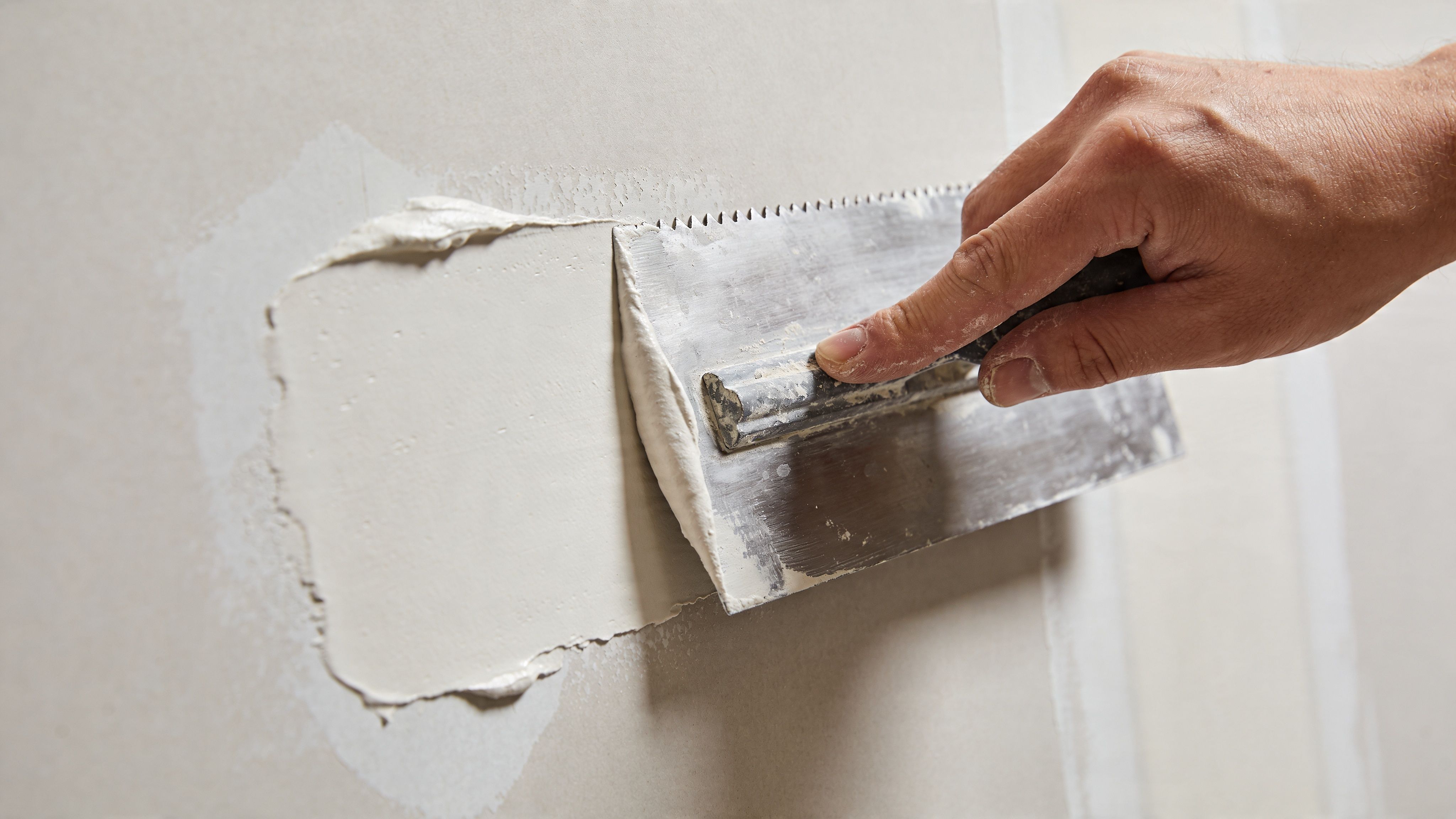 A close-up view of a hand using a metal trowel to apply joint compound on drywall.