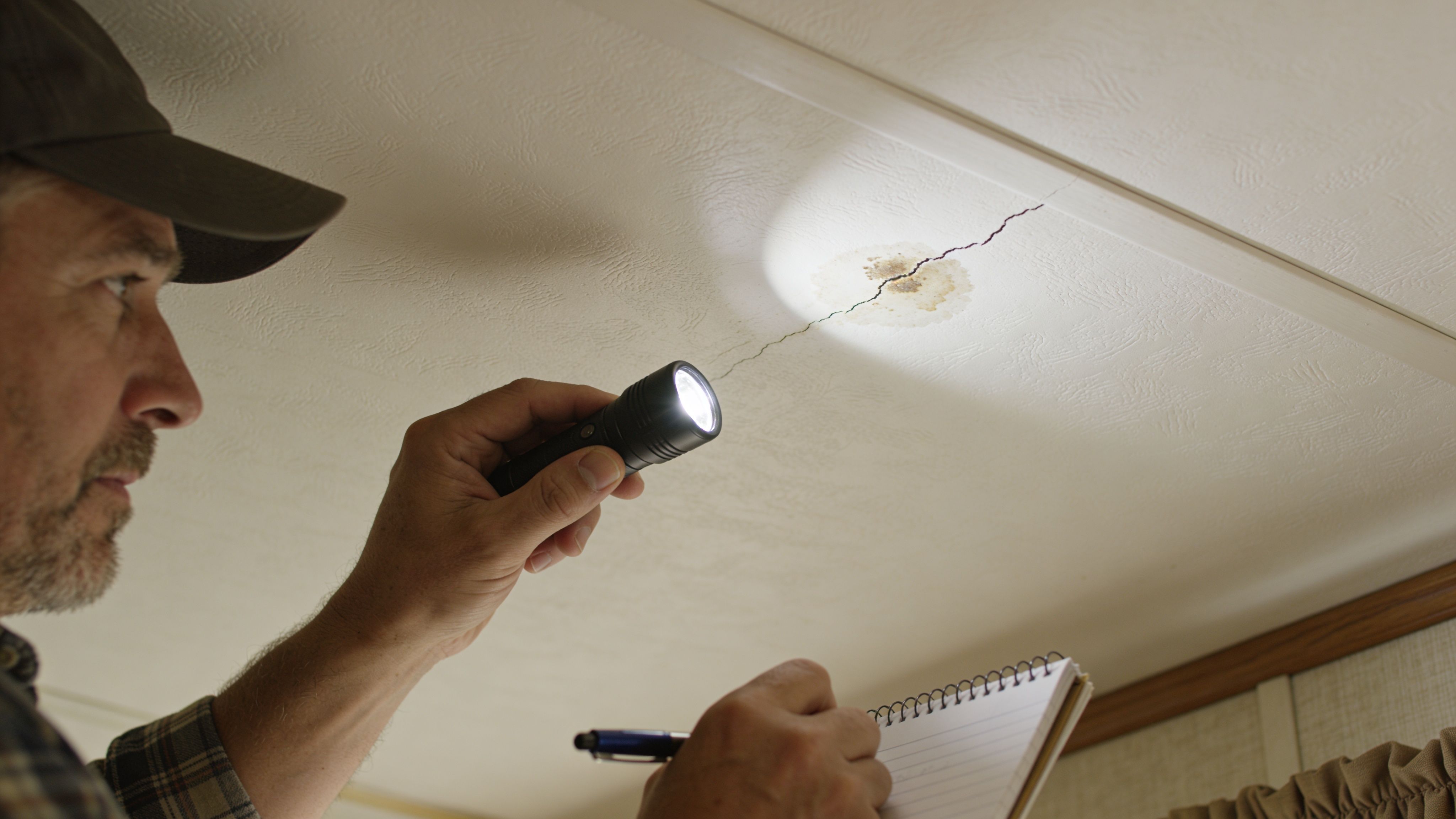 A professional building inspector examining a water-damaged crack in a mobile home ceiling with a flashlight.