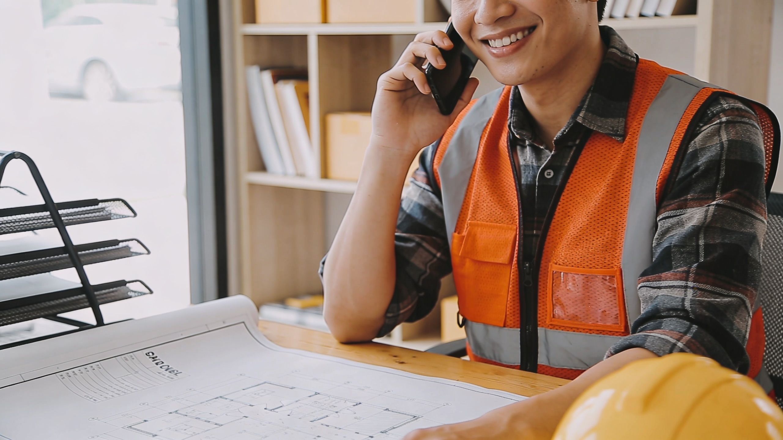 A smiling construction manager in a safety vest talking on his phone over blueprints at a desk.