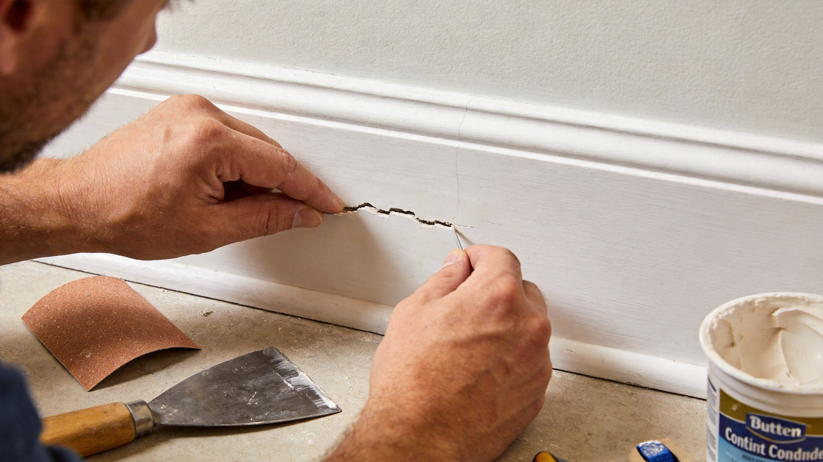 A person uses a putty knife to repair a crack in a white wooden baseboard trim.