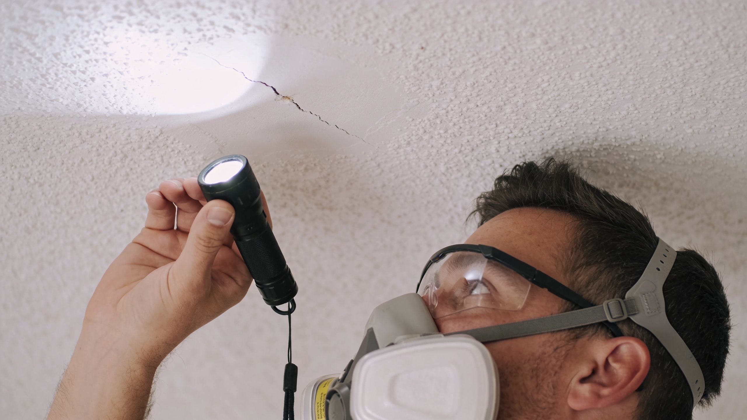 A professional inspector wearing a respirator and safety goggles examines a crack in a textured ceiling with a flashlight.