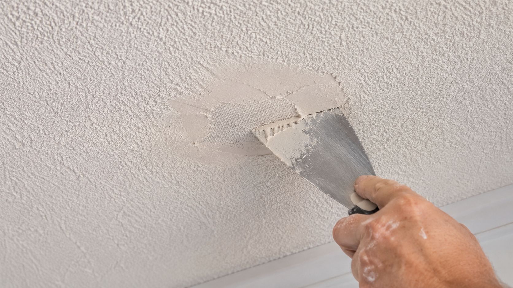 A person uses a putty knife to apply compound over a mesh patch on a textured ceiling.