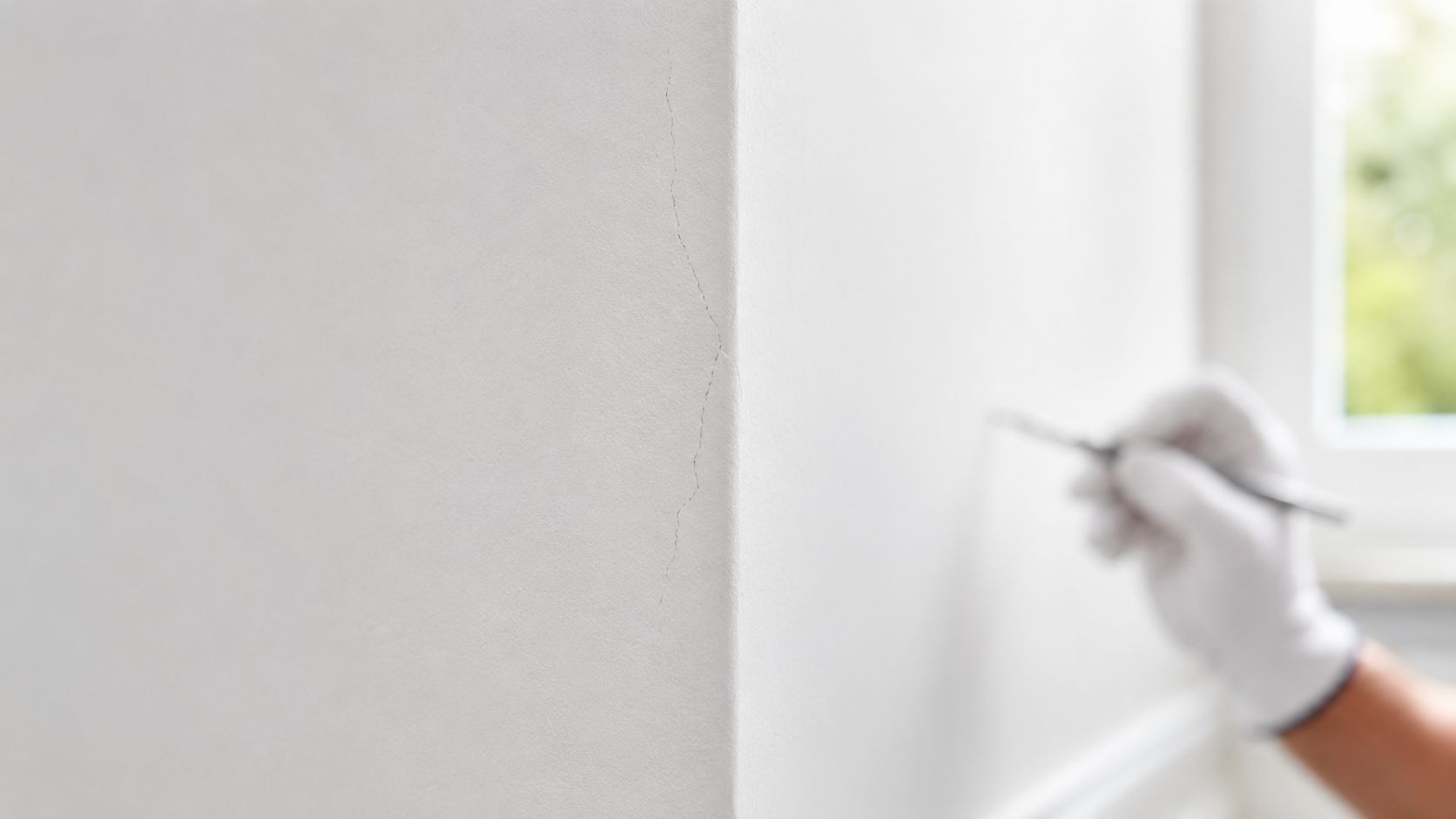 A gloved hand uses a small tool to repair a vertical crack on a white wall corner.