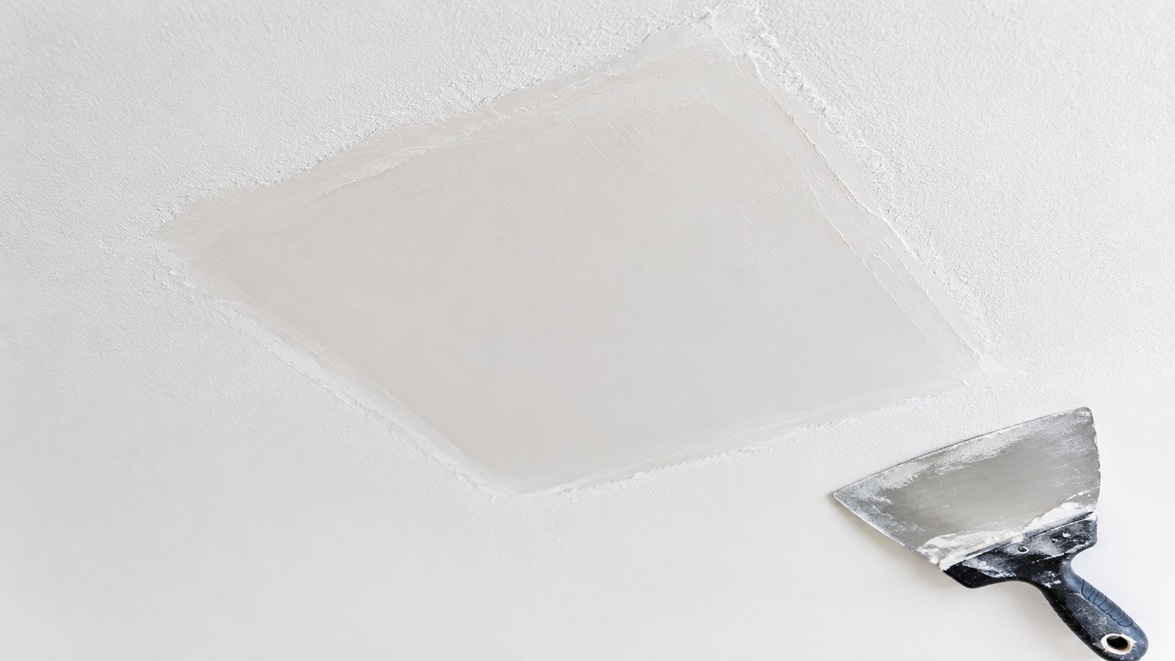 A freshly patched section of a white ceiling with a metal putty knife resting nearby.