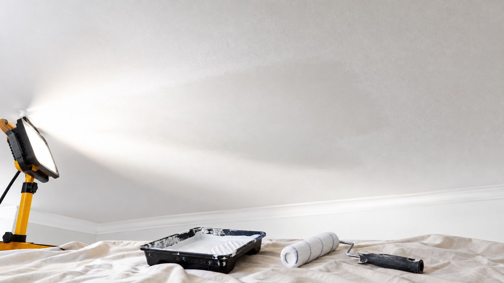 A professional work light illuminates a freshly painted white ceiling with paint tray and roller nearby.